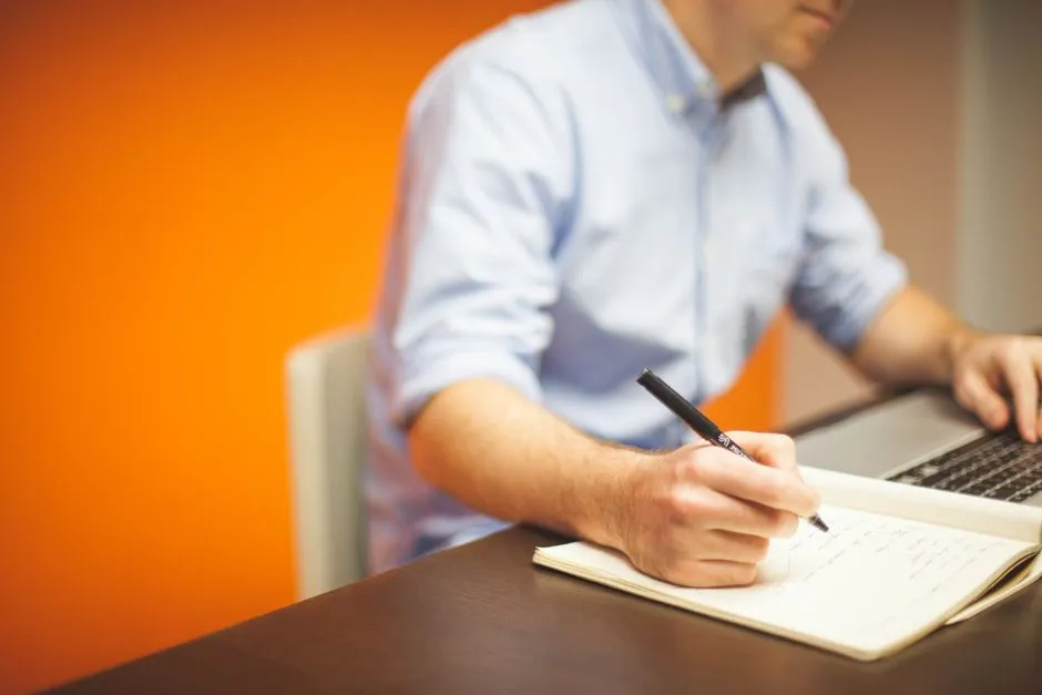 Writer taking notes at a laptop beside an open reference book